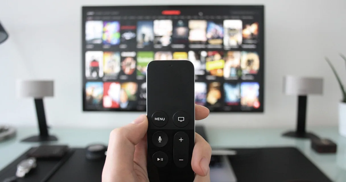 Mão segurando um controle remoto diante de uma TV com catálogo de programas na tela
