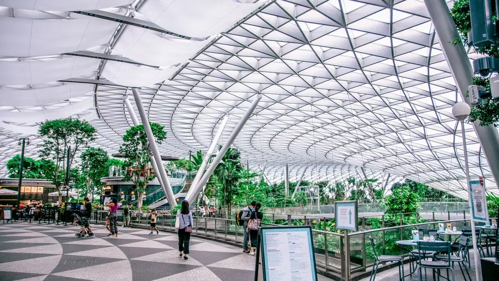 Saguão de aeroporto com teto de vidro em grade, jardim interno e passageiros caminhando